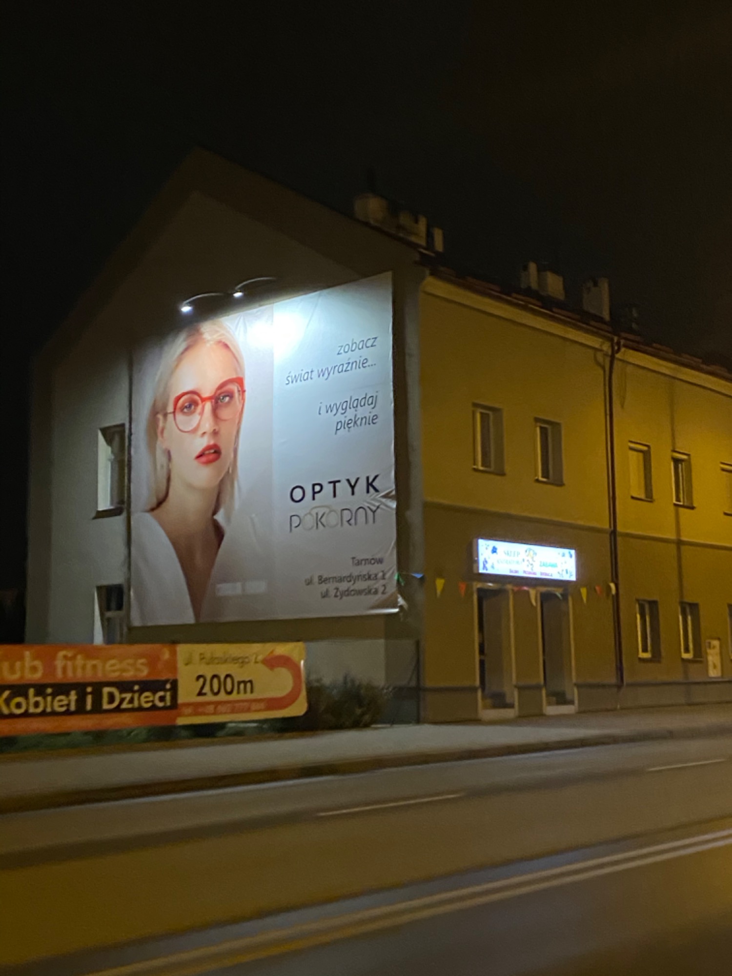 Billboard reklamowy na ścianie budynku przedstawiający kobietę w okularach z hasłem 'zobacz świat wyraźnie... i wyglądaj pięknie', reklama optyka Pokorny w Tarnowie, ujęcie nocne.