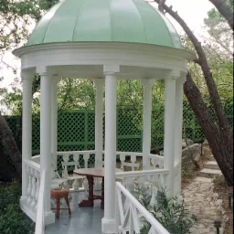 Biała altana z zielonym dachem kopułowym, otoczona drzewami i zielenią, z drewnianym stołem i krzesłami w środku, widok z zewnątrz na ścieżkę prowadzącą do altany.