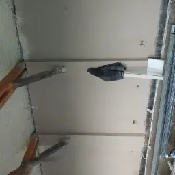 Wnętrze w trakcie remontu: gładka, jasna ściana po nałożeniu tynku, widoczne elementy instalacji elektrycznej, profile metalowe przy ścianie, drewniane podpory i pianka izolacyjna przy suficie.