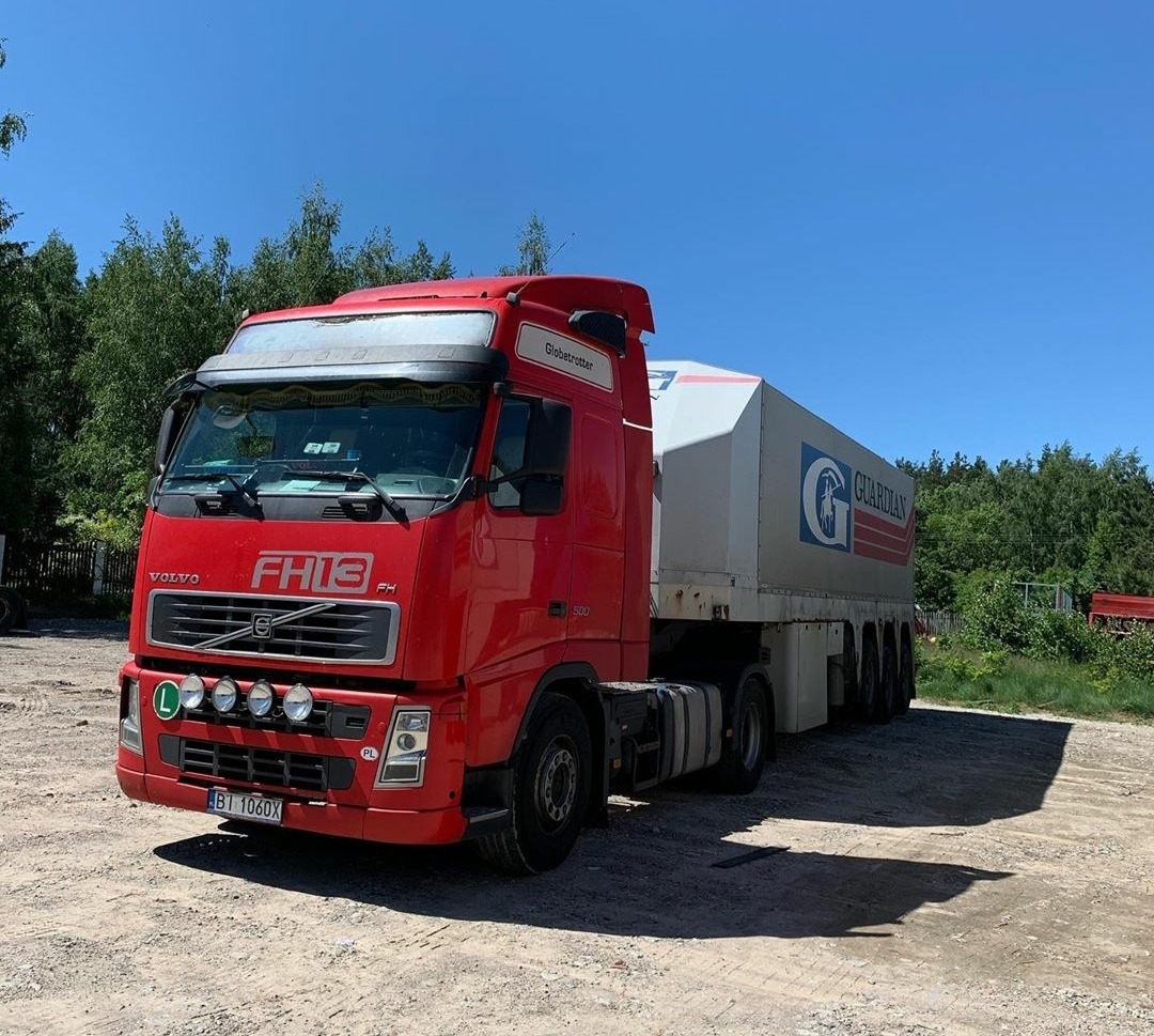 Czerwony ciągnik siodłowy Volvo FH13 z naczepą Guardian na żwirowym placu, widok z przodu, błękitne niebo w tle.