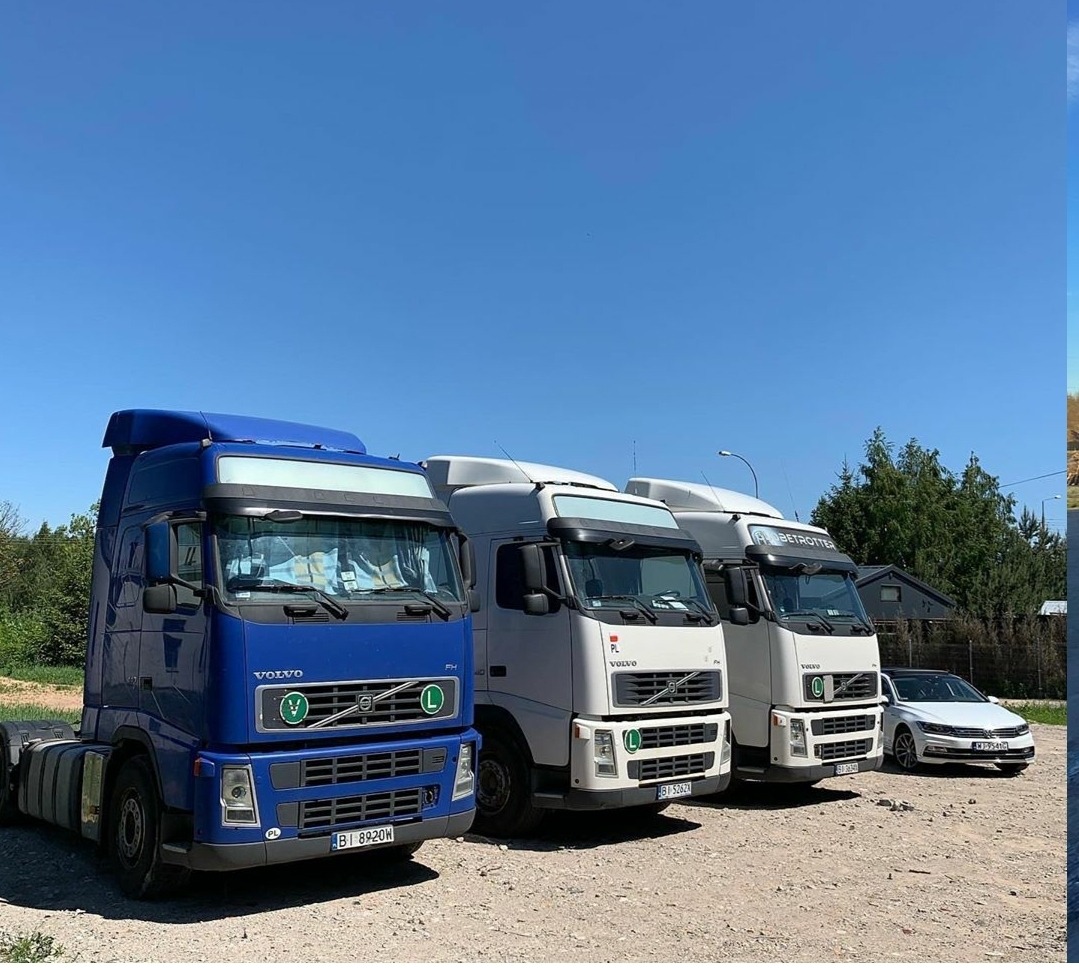 Rząd trzech ciągników siodłowych Volvo, niebieski i dwa białe, zaparkowane na żwirowym placu w słoneczny dzień.