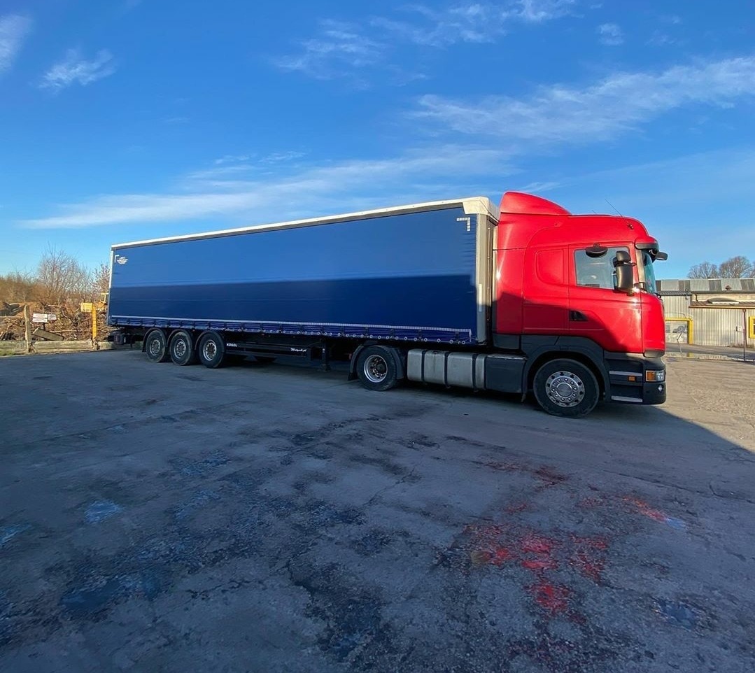 Czerwona ciężarówka Scania z niebieską naczepą na betonowym placu w słoneczny dzień.