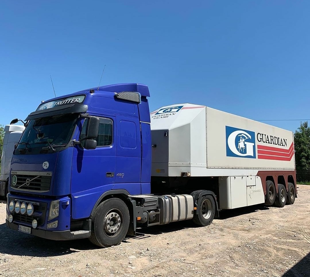 Niebieski ciągnik siodłowy Volvo z naczepą firmy Guardian Glass na żwirowym placu, widoczna tablica rejestracyjna PL.