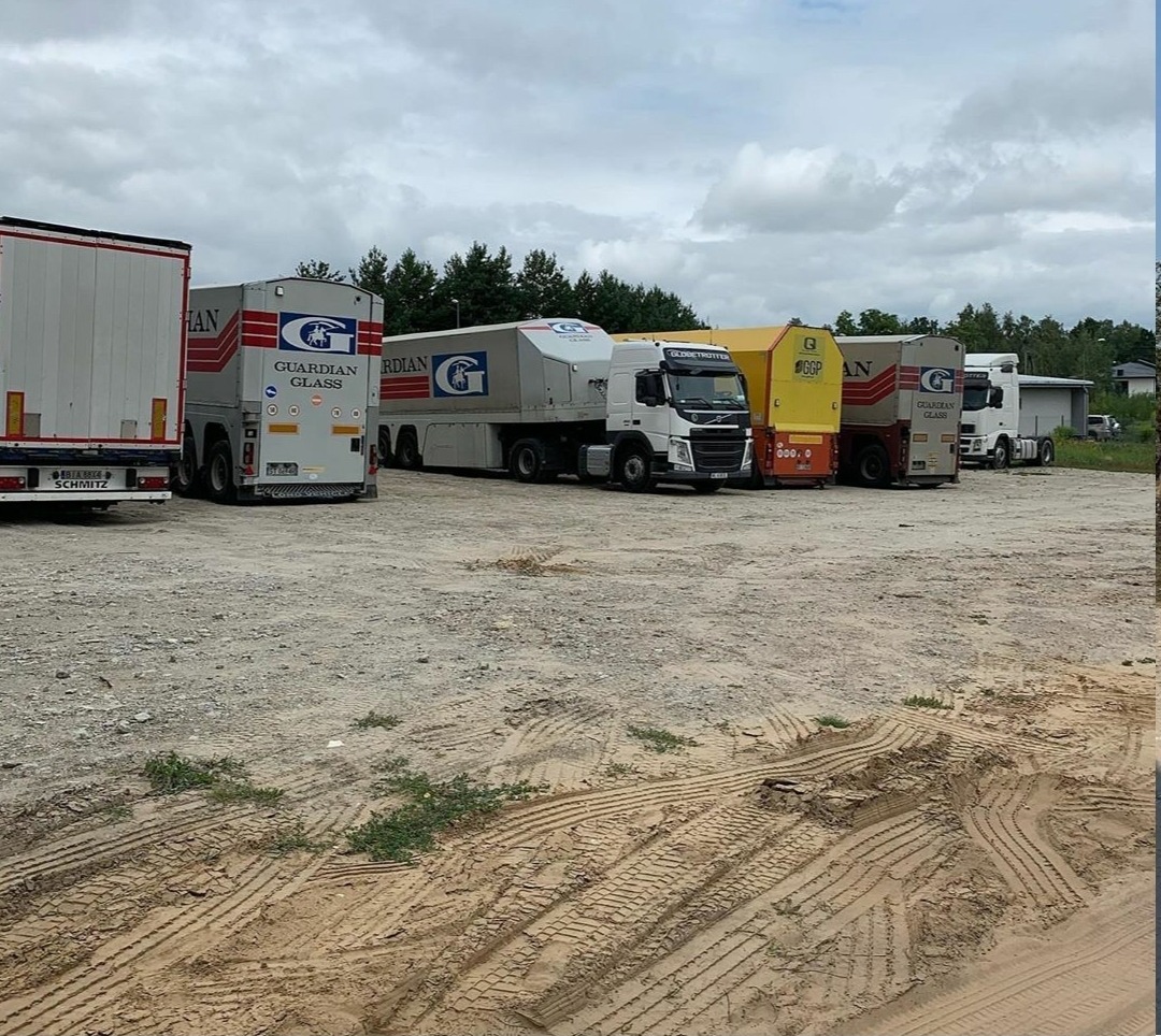 Rząd zaparkowanych ciężarówek z naczepami na żwirowym placu, z logo Guardian Glass na niektórych z nich, pod pochmurnym niebem.