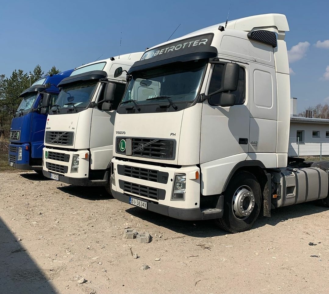 Trzy ciągniki siodłowe Volvo FH, w kolorach niebieskim i białym, stojące w rzędzie na żwirowym placu w słoneczny dzień.