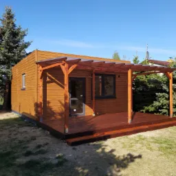 Drewniana altana z zadaszeniem pergolowym i tarasem, otoczona zielenią w słoneczny dzień.