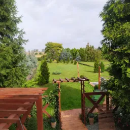 Widok na zadbany ogród z drewnianym tarasem i pergolą, z drewnianą ścieżką i leżakiem; roślinność różnorodna, w tle pole uprawne.