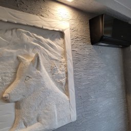 Cold - Man Patryk Kozera - Czarny klimatyzator ścienny marki AUX zamontowany nad płaskorzeźbą z motywem wilka na teksturowanej, szarej ścianie.