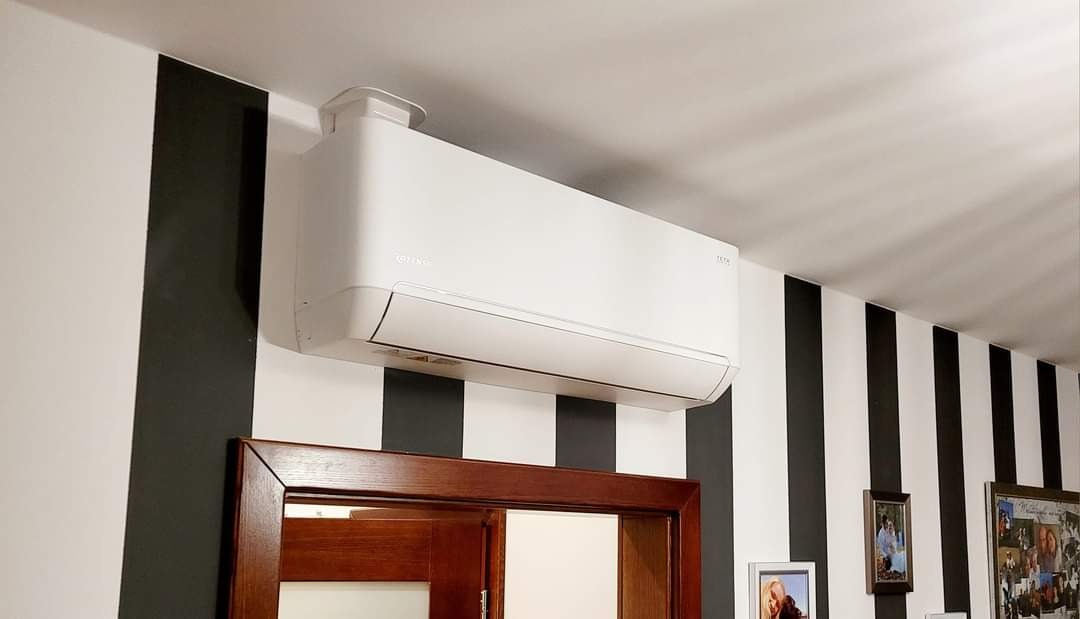 Biały klimatyzator marki Tesla zamontowany na ścianie w czarno-białe pasy nad drewnianymi drzwiami.