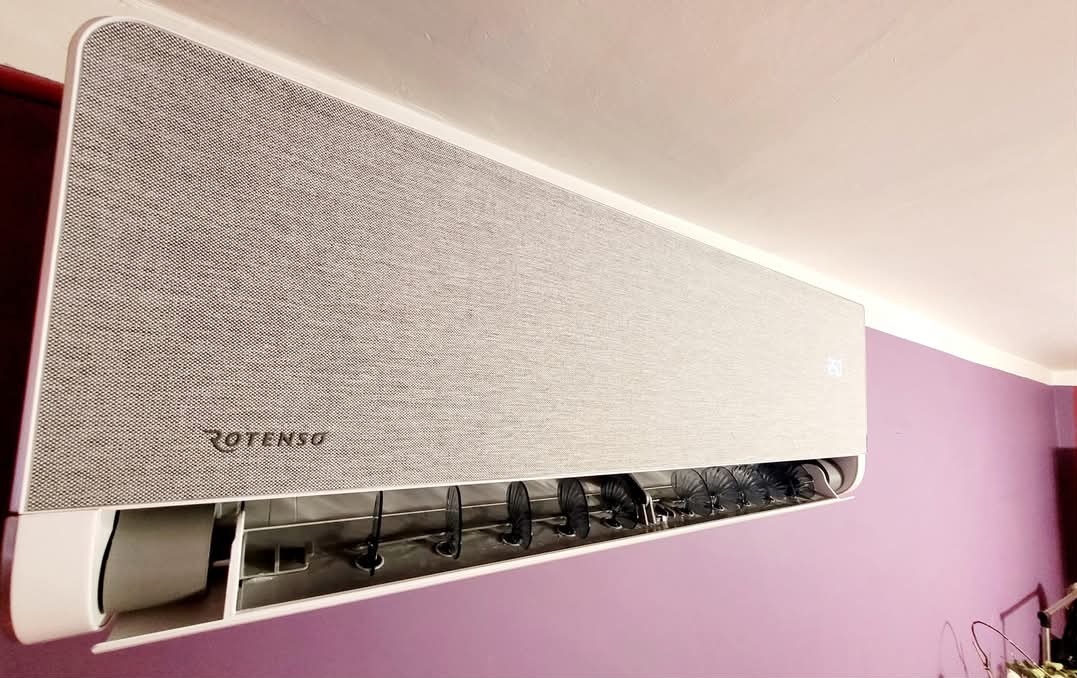 Szary klimatyzator Rotenso z widocznymi żaluzjami, zamontowany na fioletowej ścianie w pomieszczeniu.