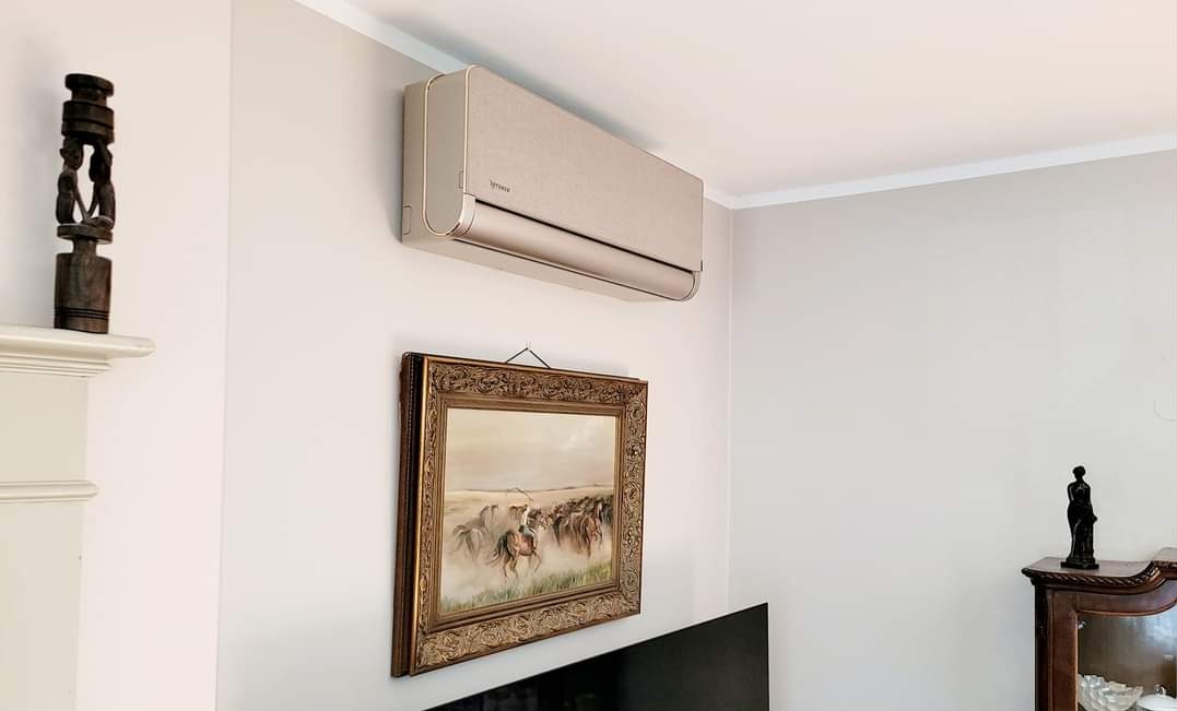 Beżowa klimatyzacja ścienna zamontowana w salonie, widoczna nad obrazem w złotej ramie i fragmentem telewizora.