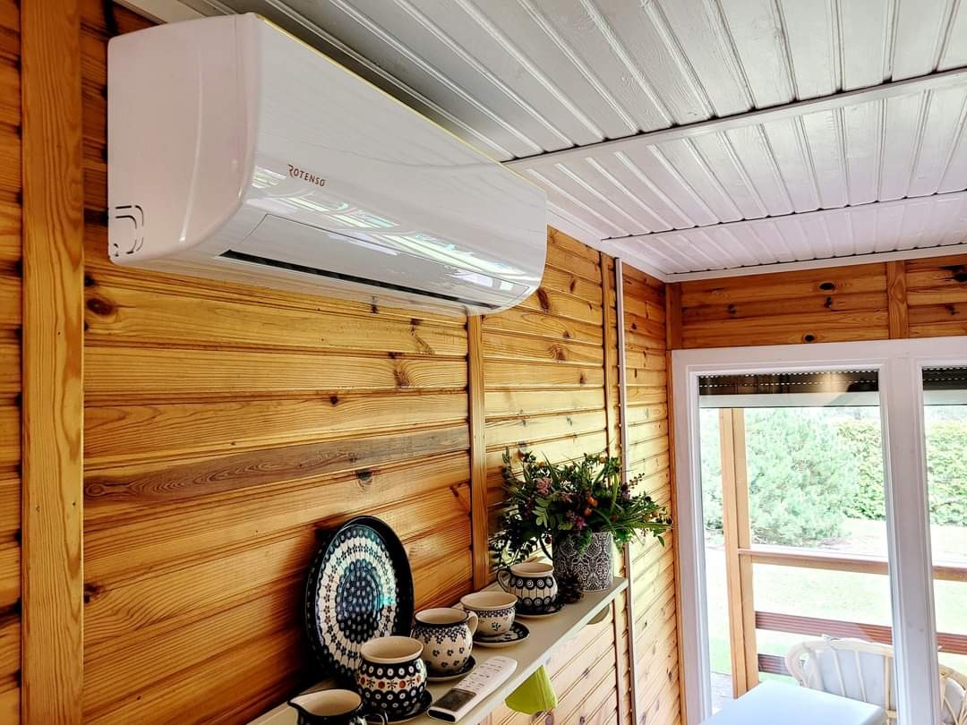 Biały klimatyzator marki Rotenso zamontowany na drewnianej ścianie w domowym wnętrzu w Słubicach, widok na okno i dekoracyjne naczynia na półce.