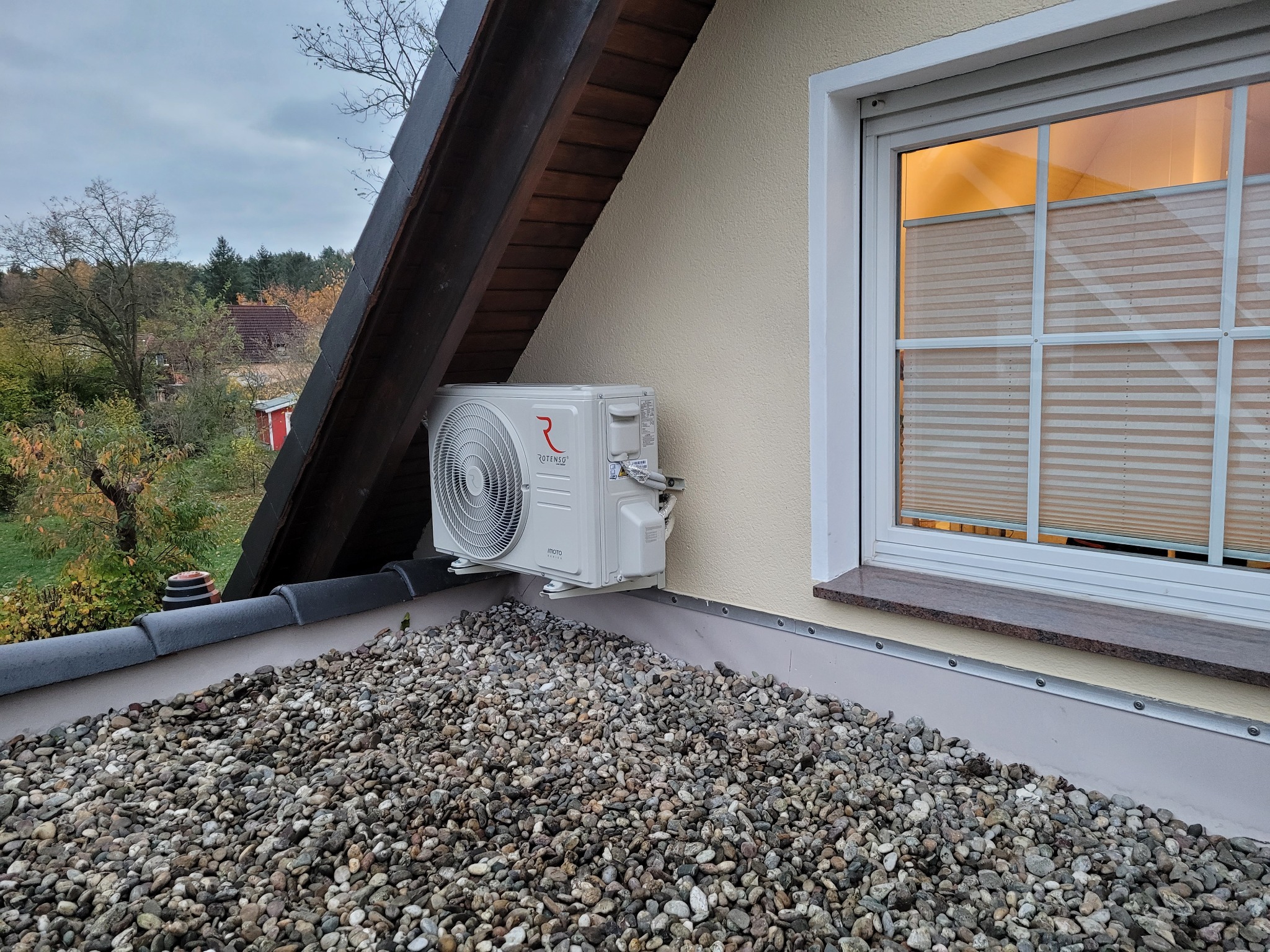 Biała jednostka zewnętrzna klimatyzacji Rotenso zamontowana na dachu pokrytym kamieniami, widok z boku na tle okna z żaluzjami plisowanymi i drewnianej konstrukcji dachu.