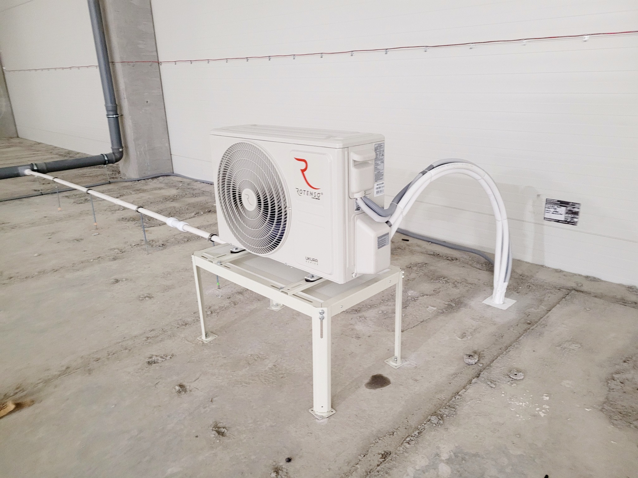 Biała jednostka zewnętrzna klimatyzacji Rotenso zamontowana na metalowym stelażu w surowym wnętrzu hali, widoczne rury instalacyjne i betonowa podłoga w trakcie prac wykończeniowych.
