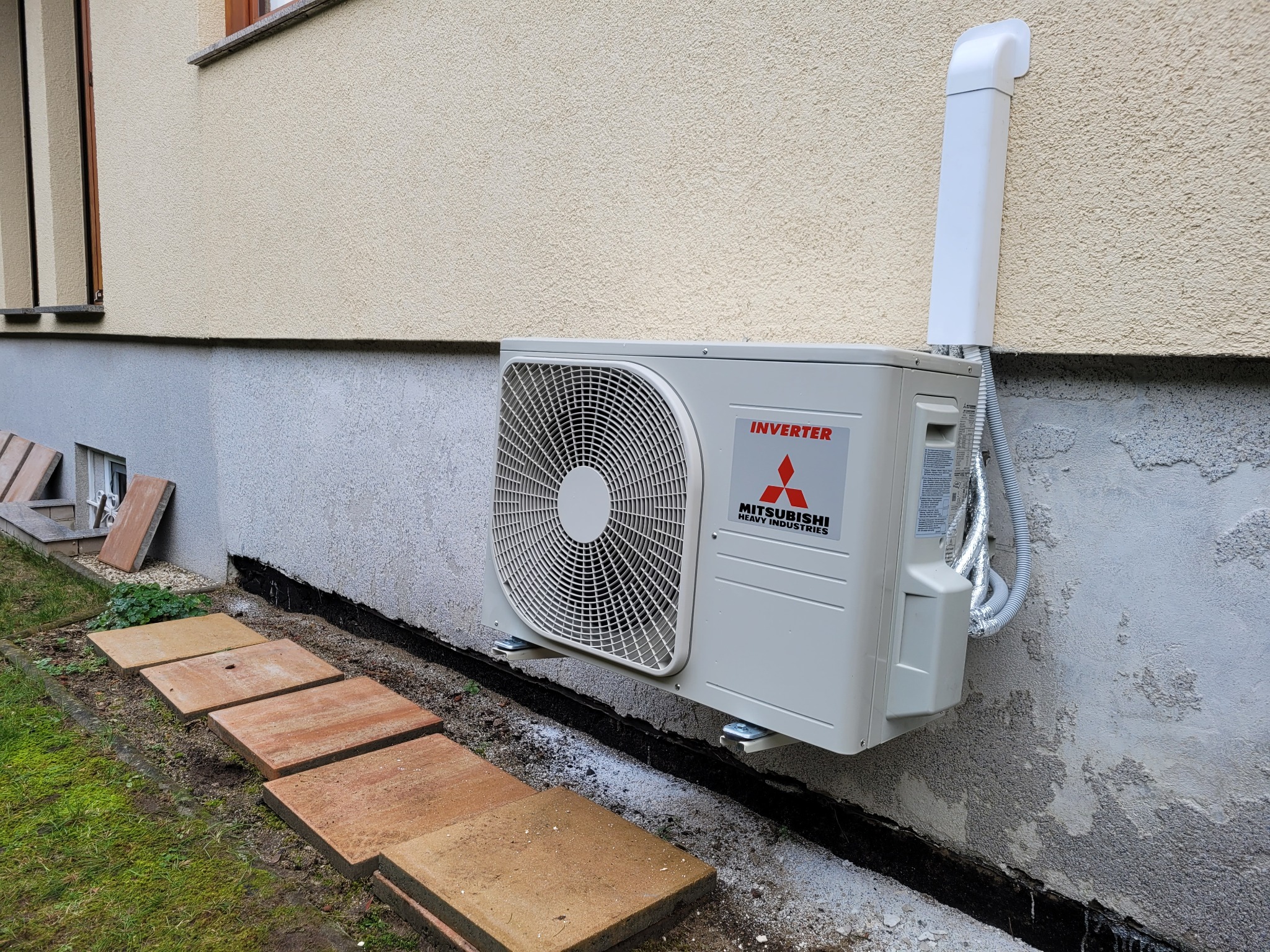 Zewnętrzna jednostka klimatyzacji Mitsubishi Heavy Industries zamontowana na ścianie budynku z widocznymi przewodami w osłonie i fragmentem chodnika z płyt.