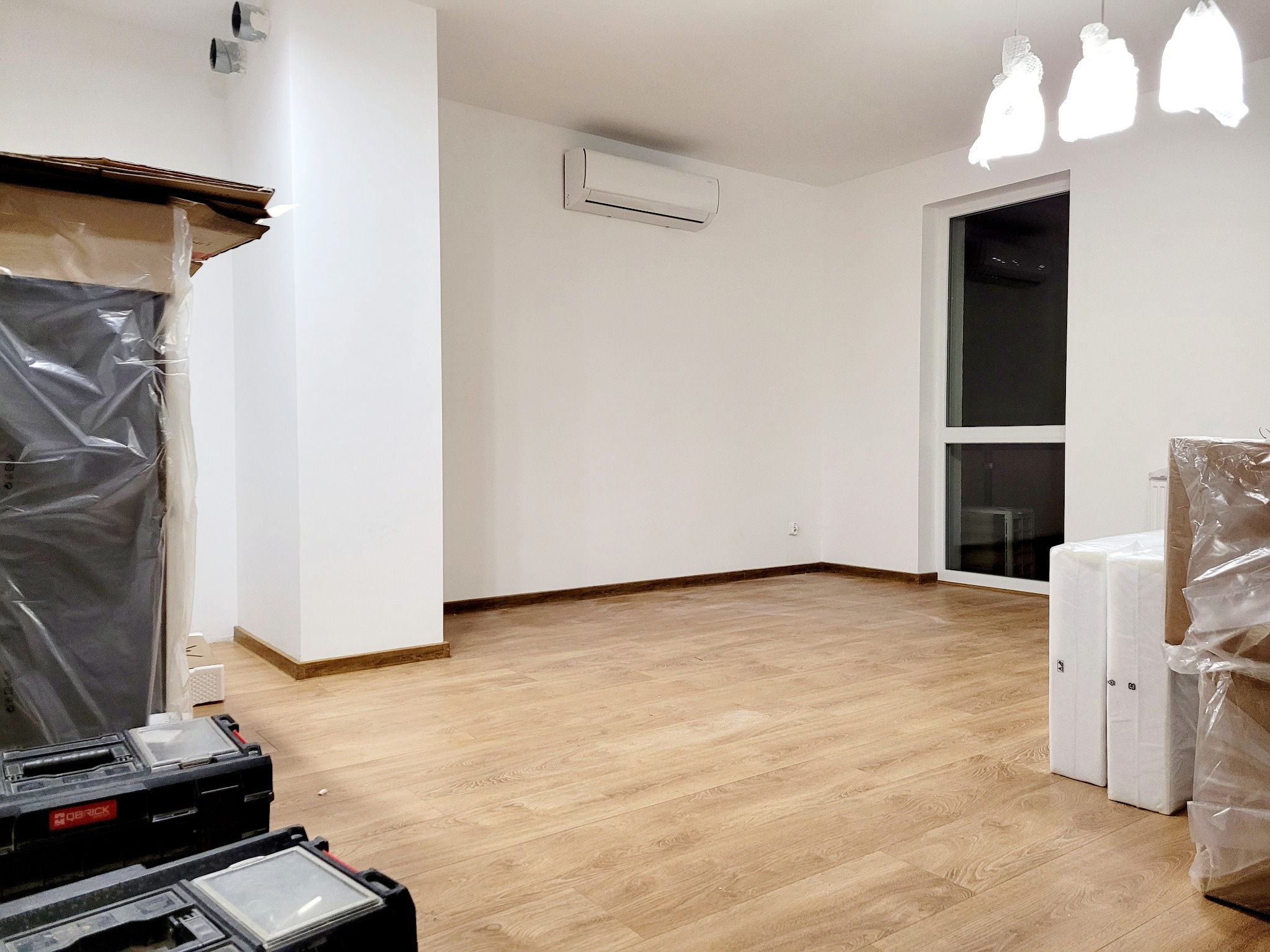 Białe wnętrze z zamontowaną klimatyzacją na ścianie, podłoga wyłożona panelami, widoczne narzędzia i elementy wyposażenia w folii ochronnej.