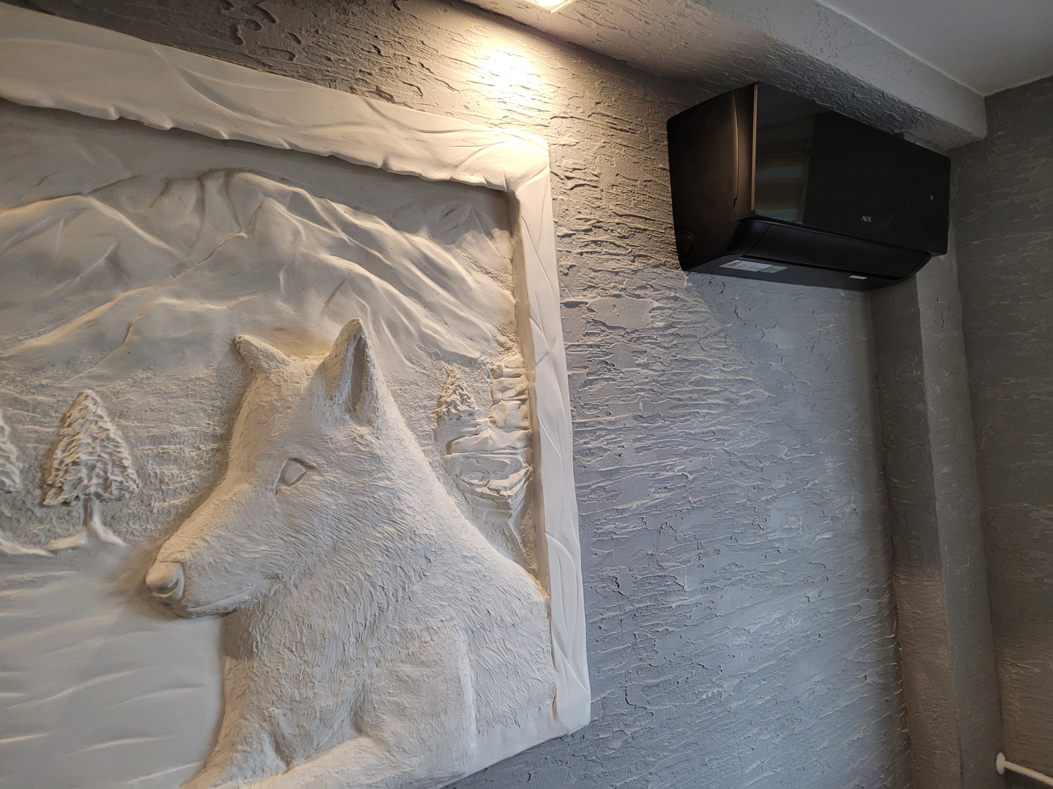 Czarny klimatyzator ścienny marki AUX zamontowany nad płaskorzeźbą z motywem wilka na teksturowanej, szarej ścianie.