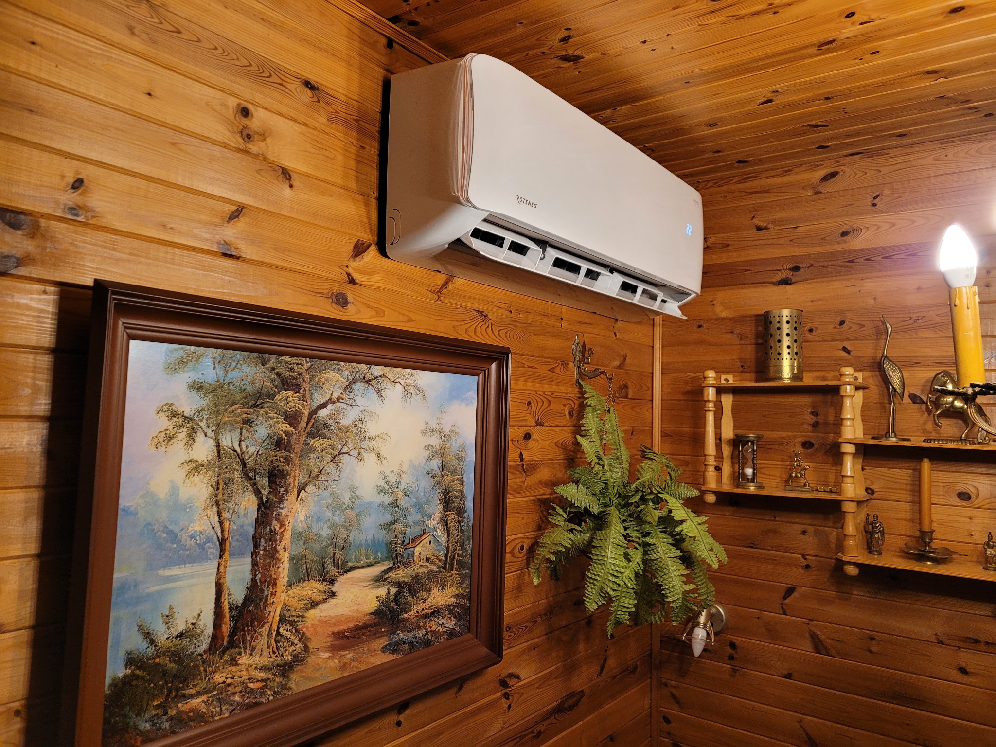 Biały klimatyzator marki Rotenso zamontowany na ścianie z boazerii, obok wisi obraz z pejzażem i półka z ozdobami.