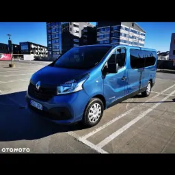 Niebieski bus Renault Trafic zaparkowany na parkingu z widokiem na nowoczesny budynek biurowy w tle.