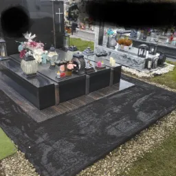 Czarny, podwójny pomnik z granitu z kwiatami i lampionami, otoczony czarną kostką brukową, drobnymi kamieniami i sztuczną trawą.