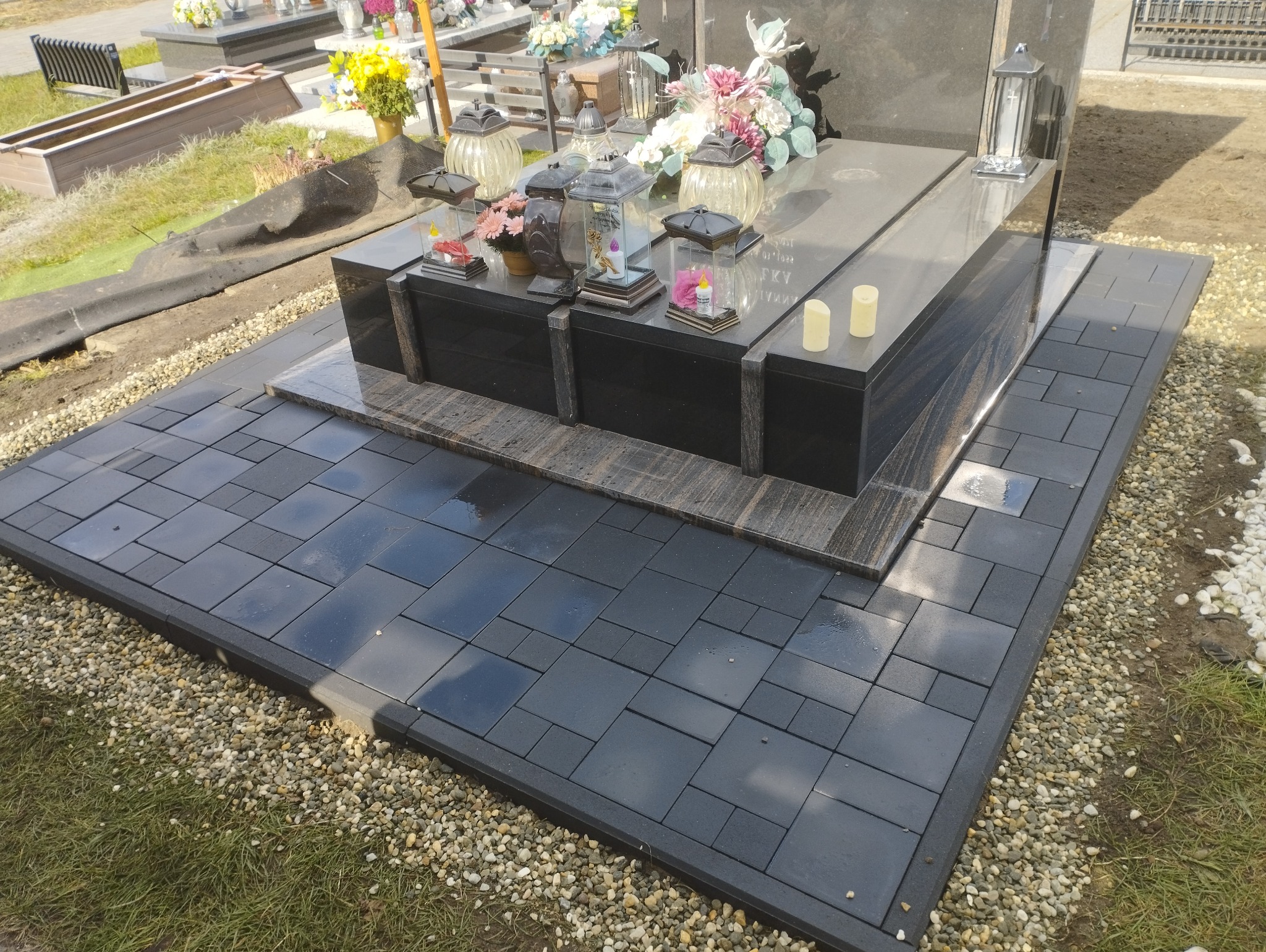 Czarny nagrobek z ciemnymi lampionami i kwiatami, otoczony świeżo ułożoną, błyszczącą kostką brukową w szarym odcieniu, wykończoną drobnym żwirem.