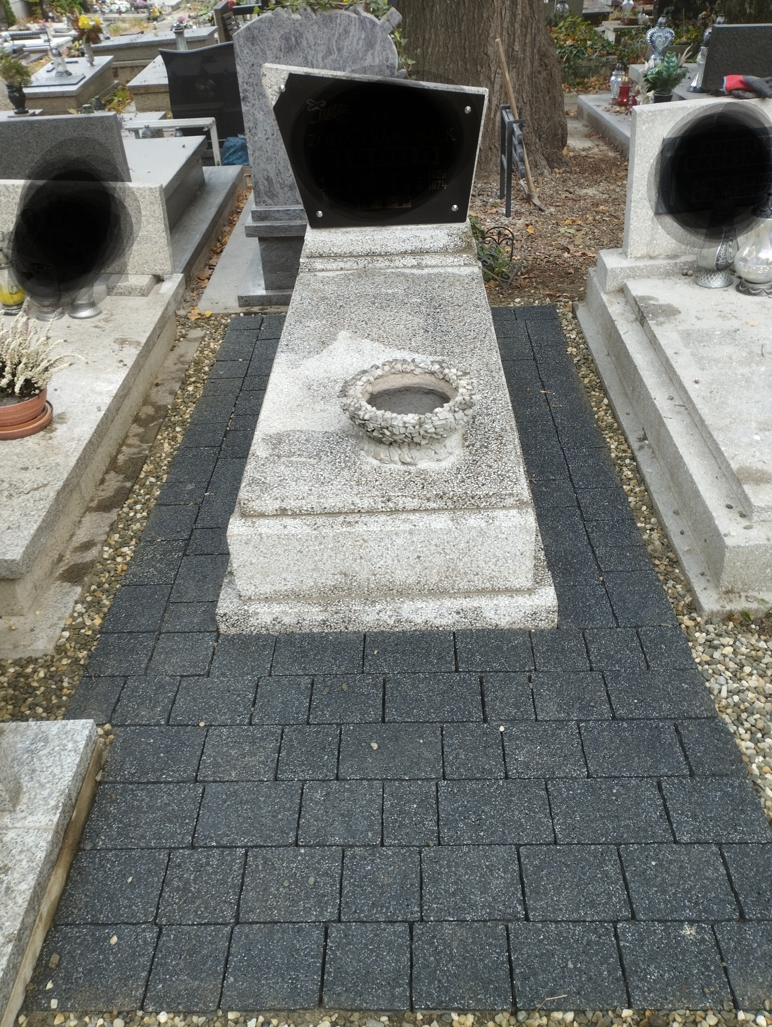 Ułożona czarna kostka brukowa prowadząca do nagrobka z kamiennym wieńcem, otoczona drobnymi kamykami.