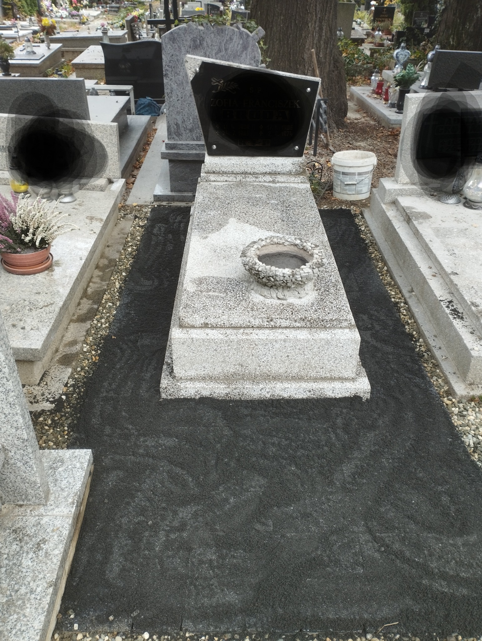 Granitowy nagrobek z czarną tablicą, otoczony świeżo rozsypanym, ciemnym grysem. Na płycie nagrobnej kamienna misa na kwiaty.