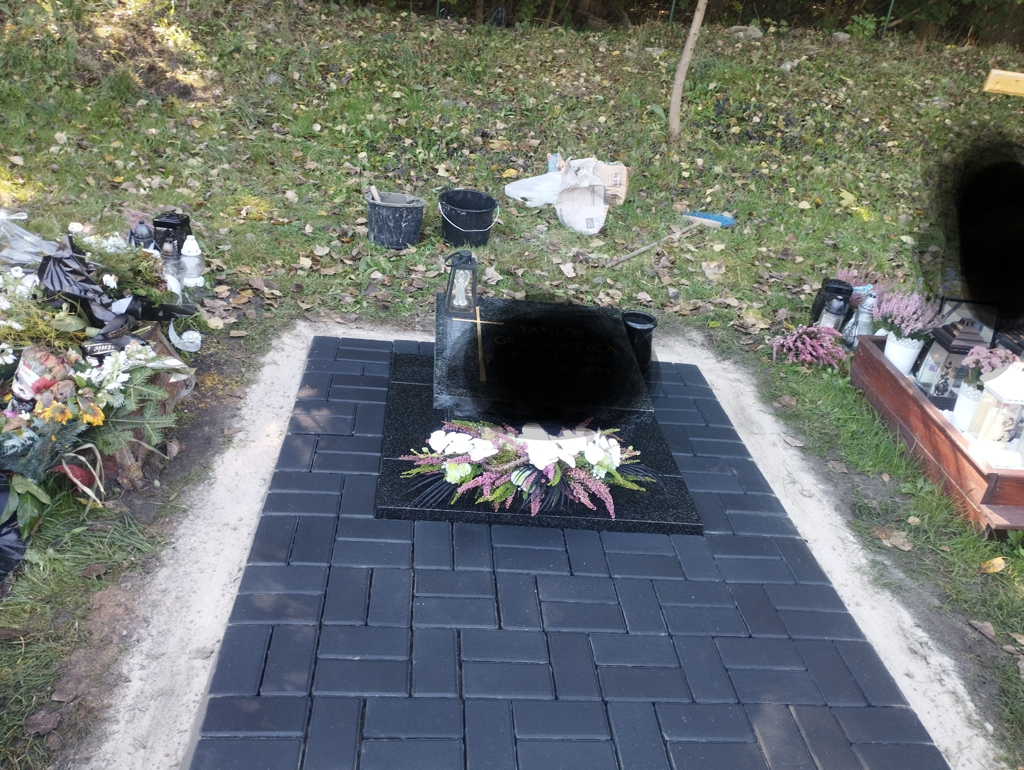 Czarny nagrobek z krzyżem i kwiatami, otoczony świeżo ułożoną, ciemną kostką brukową na cmentarzu, widoczne ślady prac ziemnych i narzędzia.