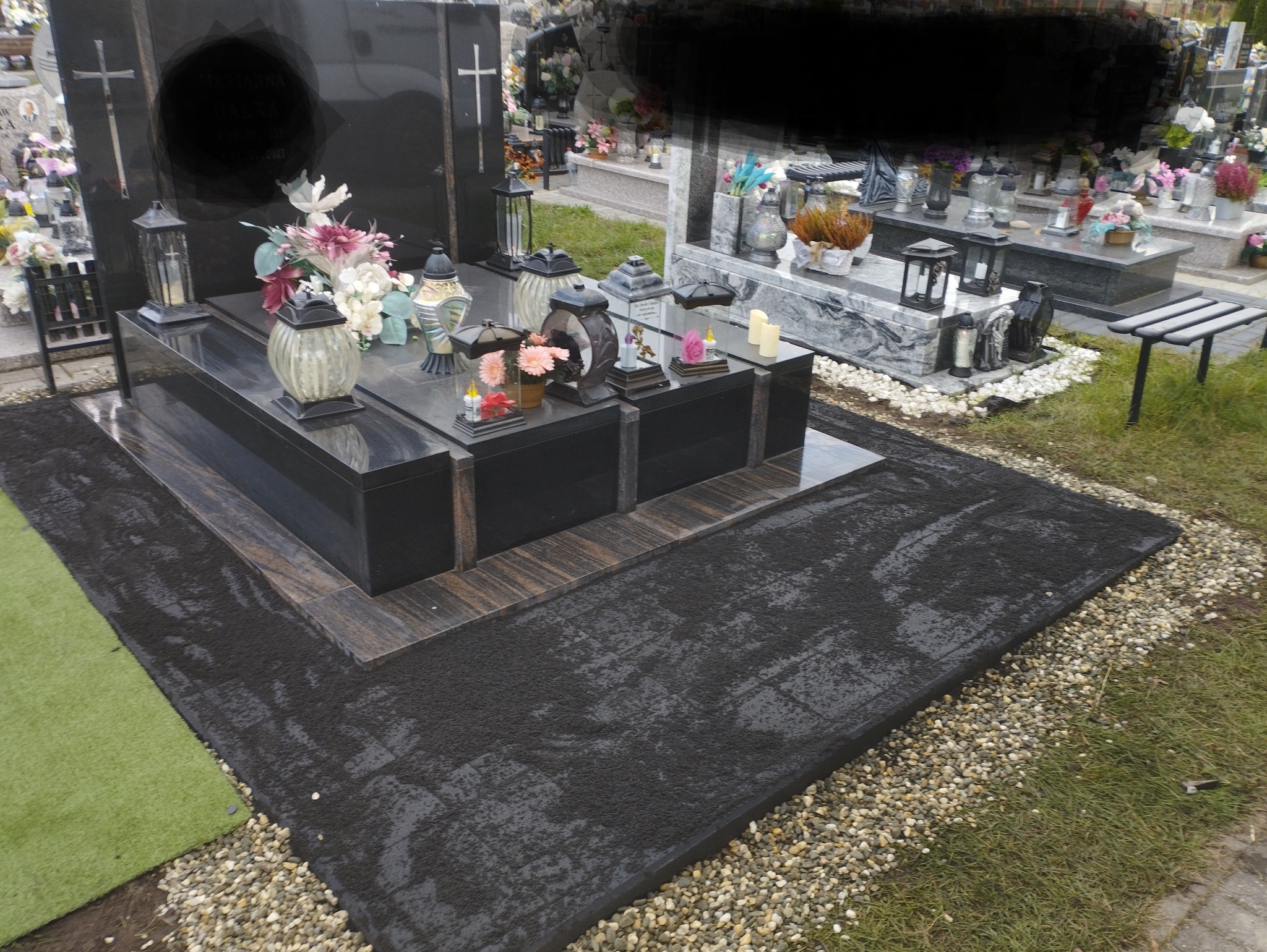 Czarny, podwójny pomnik z granitu z kwiatami i lampionami, otoczony czarną kostką brukową, drobnymi kamieniami i sztuczną trawą.