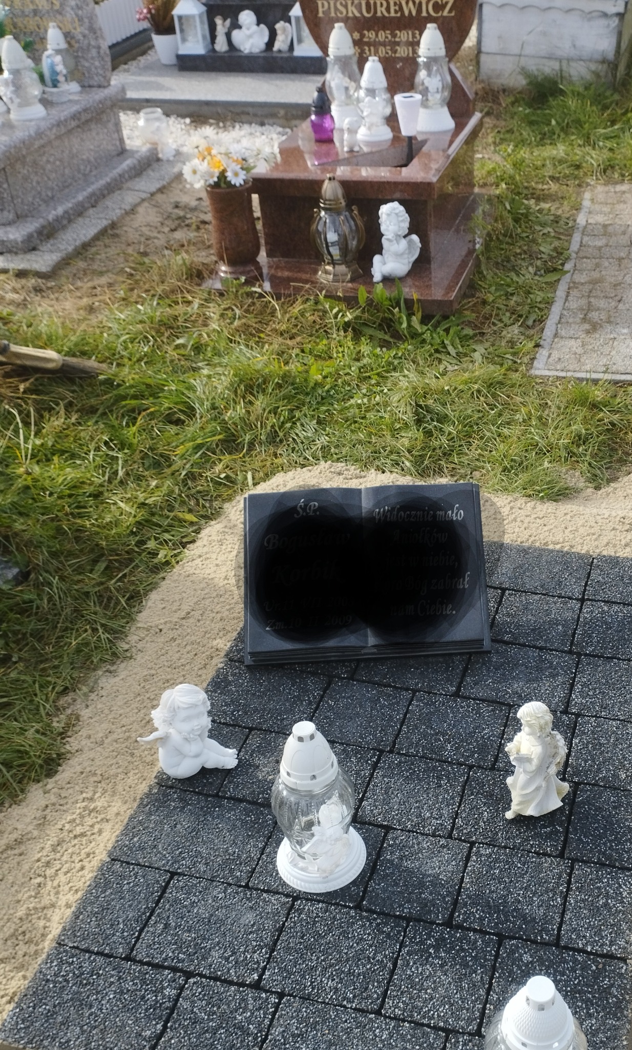 Nagrobek w formie otwartej księgi z wyrytym tekstem, otoczony szarą kostką brukową, lampionami i figurkami aniołków na cmentarzu.