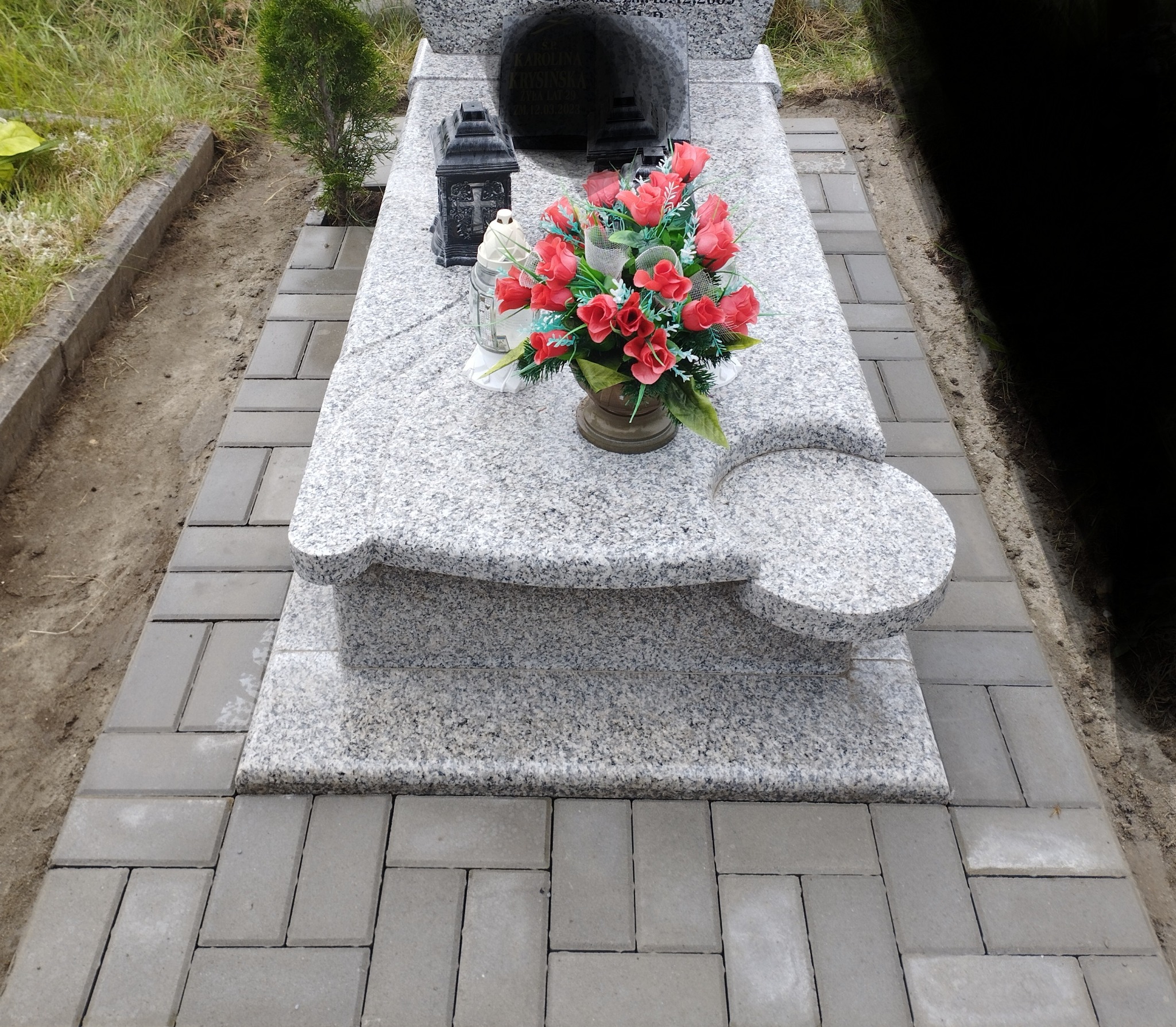 Granitowy nagrobek z kwiatami i lampionem, otoczony szarą kostką brukową