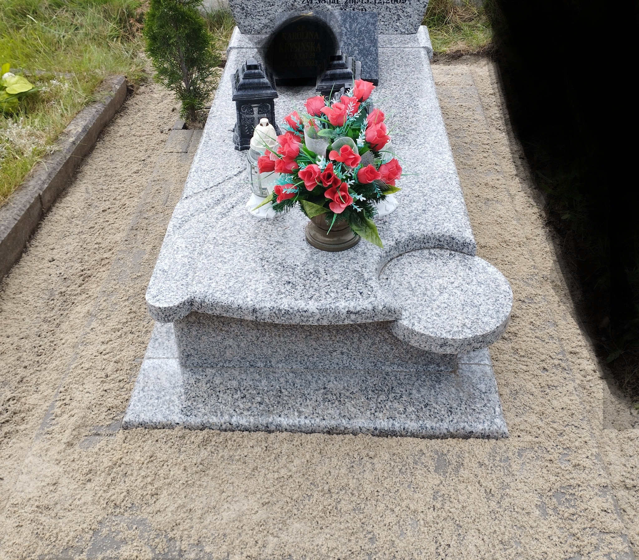 Granitowy nagrobek z wazonem czerwonych róż, lampionami i świecą, otoczony piaskiem.