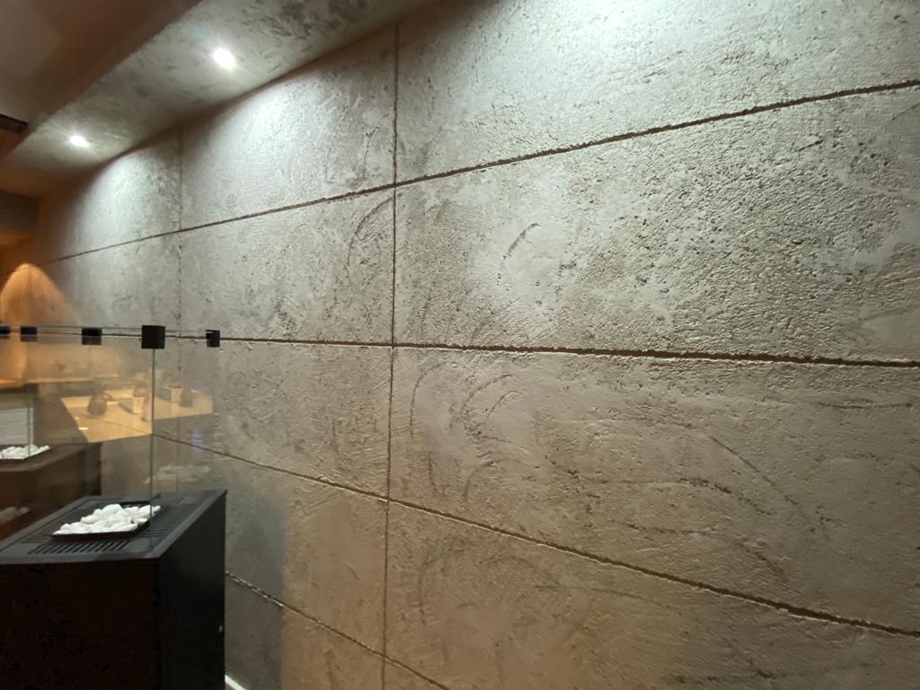 Ściana z dekoracyjną, szarą fakturą przypominającą beton, podzielona na poziome pasy, oświetlona punktowymi lampami sufitowymi. W tle widoczny fragment przeszklonej przestrzeni z elementami...