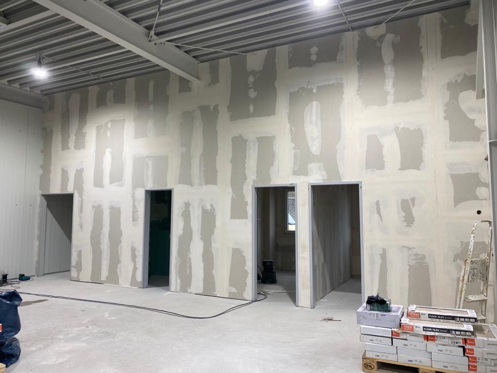 Wnętrze komercyjne w trakcie szpachlowania ścian gipsowo-kartonowych, widoczne przygotowane otwory drzwiowe, metalowa konstrukcja sufitu oraz paleta z płytkami podłogowymi.