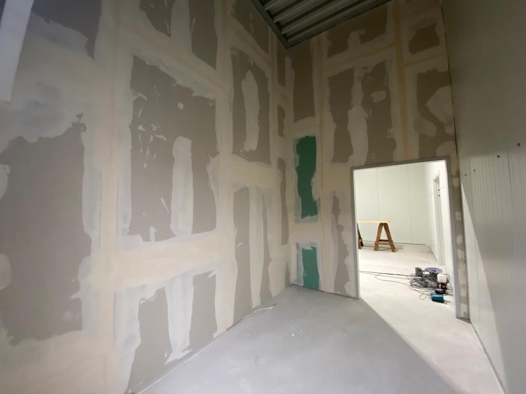 Wnętrze w trakcie remontu: ściany z płyt kartonowo-gipsowych z widocznymi łączeniami szpachlowanymi, fragmenty zielonej farby na ścianie, podłoga w szarym odcieniu, w tle widoczny drewniany kozioł...