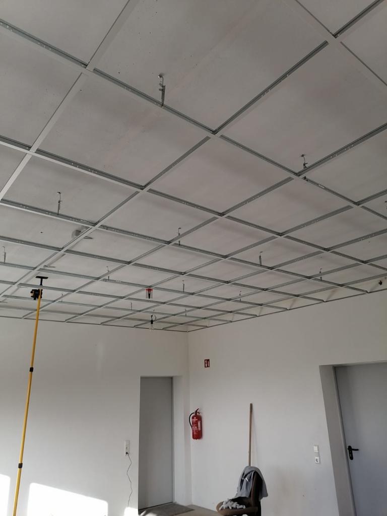 Montaż sufitu podwieszanego z widoczną metalową konstrukcją kratową i częściowo zamontowanymi panelami sufitowymi w jasnym pomieszczeniu biurowym, z widocznym miernikiem laserowym i gaśnicą...