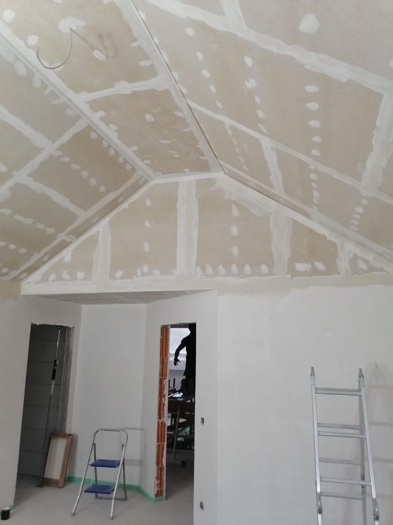 Poddasze w trakcie wykańczania, widoczne płyty gipsowo-kartonowe na skośnym suficie, zaszpachlowane łączenia, drabina oparta o ścianę, surowy stan wnętrza.