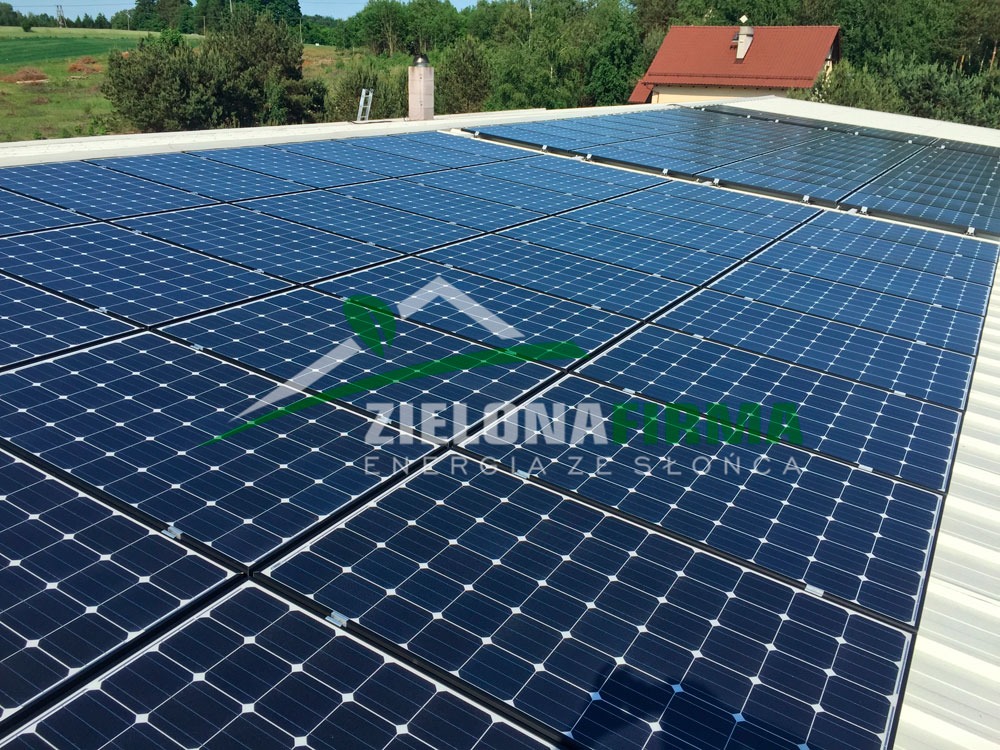 Instalacja fotowoltaiczna na dachu budynku przemysłowego w Kartuzach, widoczny komin i drabina, w tle drzewa i dom z czerwoną dachówką.