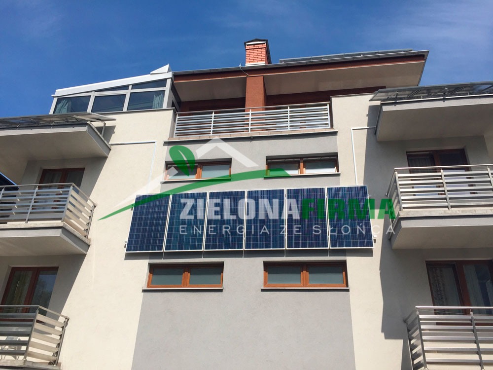 Panele fotowoltaiczne zamontowane na elewacji budynku mieszkalnego, widoczne balkony z metalowymi balustradami i logo firmy 'Zielona Energia ze Słońca' na panelach.