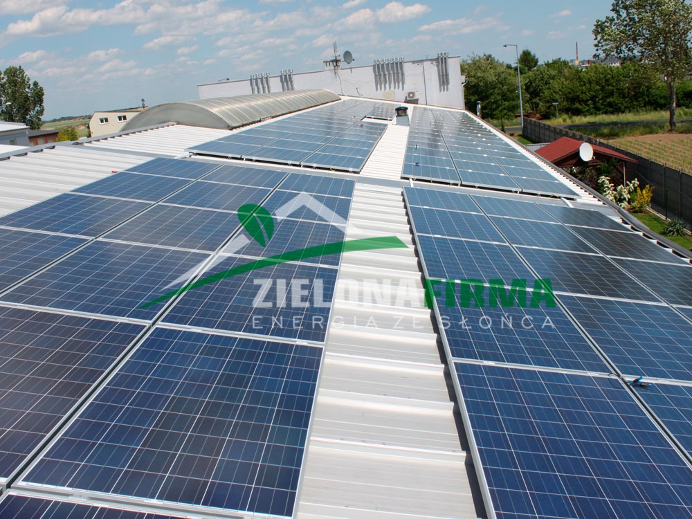 Instalacja paneli słonecznych na dachu budynku przemysłowego z widocznym logo firmy na pierwszym planie.