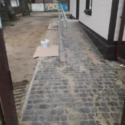 Świeżo ułożona, szara kostka brukowa prowadząca do budynku z metalową barierką, widoczne ślady piasku i wiadro z zaprawą, w tle malowidła na murze.