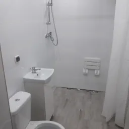 Jasna łazienka z białymi płytkami, toaletą, umywalką z szafką, prysznicem z deszczownicą i składanym siedziskiem dla osób z niepełnosprawnościami.