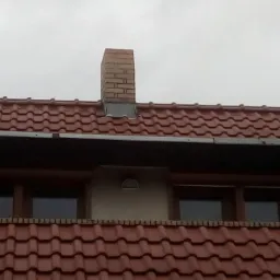 Czerwona dachówka ceramiczna z kominem z cegły, widoczne okno dachowe i rynna, pochmurne niebo w tle.