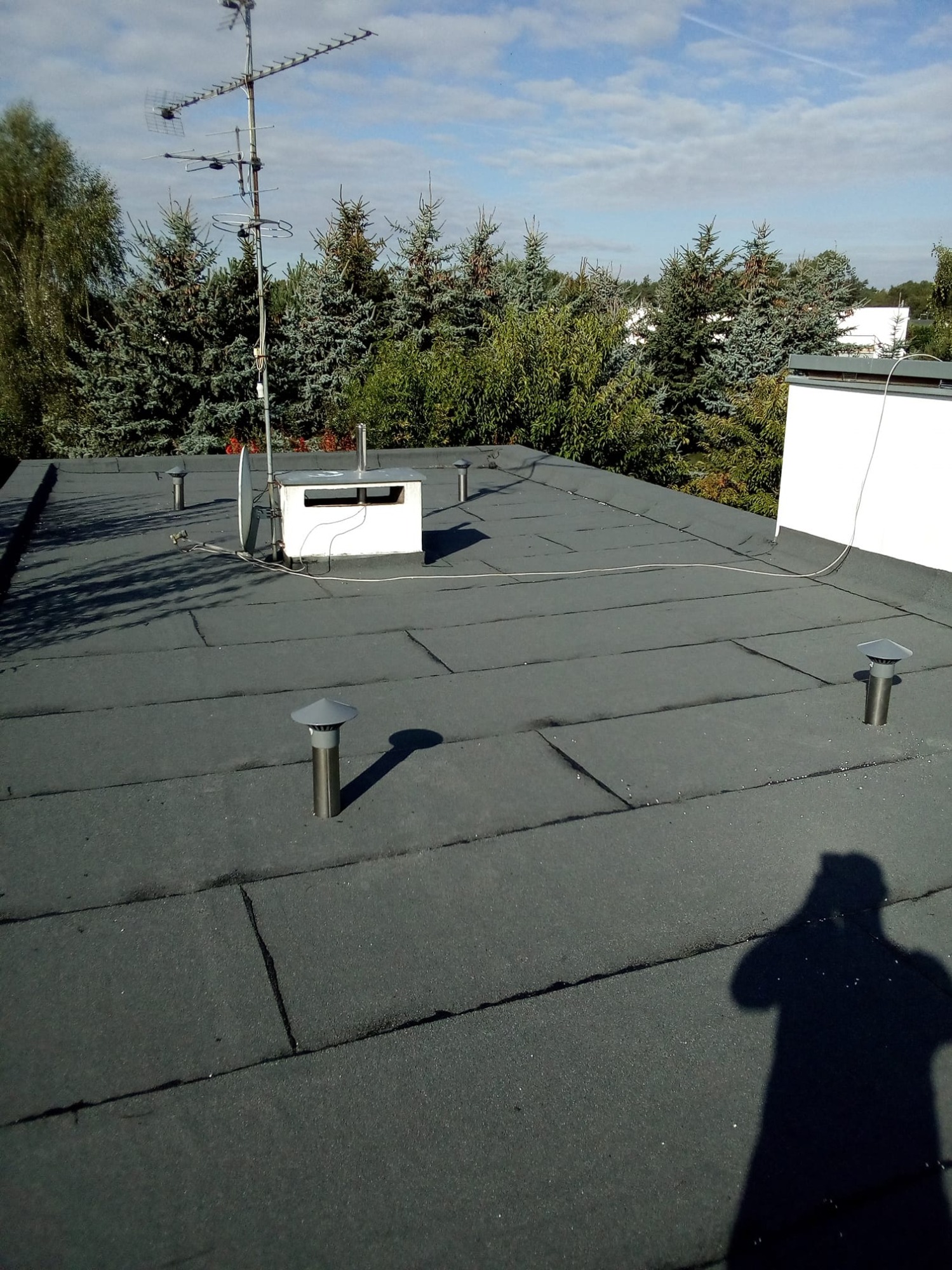 Płaski dach pokryty papą z kominkami wentylacyjnymi i anteną telewizyjną na tle zielonych drzew i błękitnego nieba z chmurami; cień fotografa widoczny w prawym dolnym rogu.
