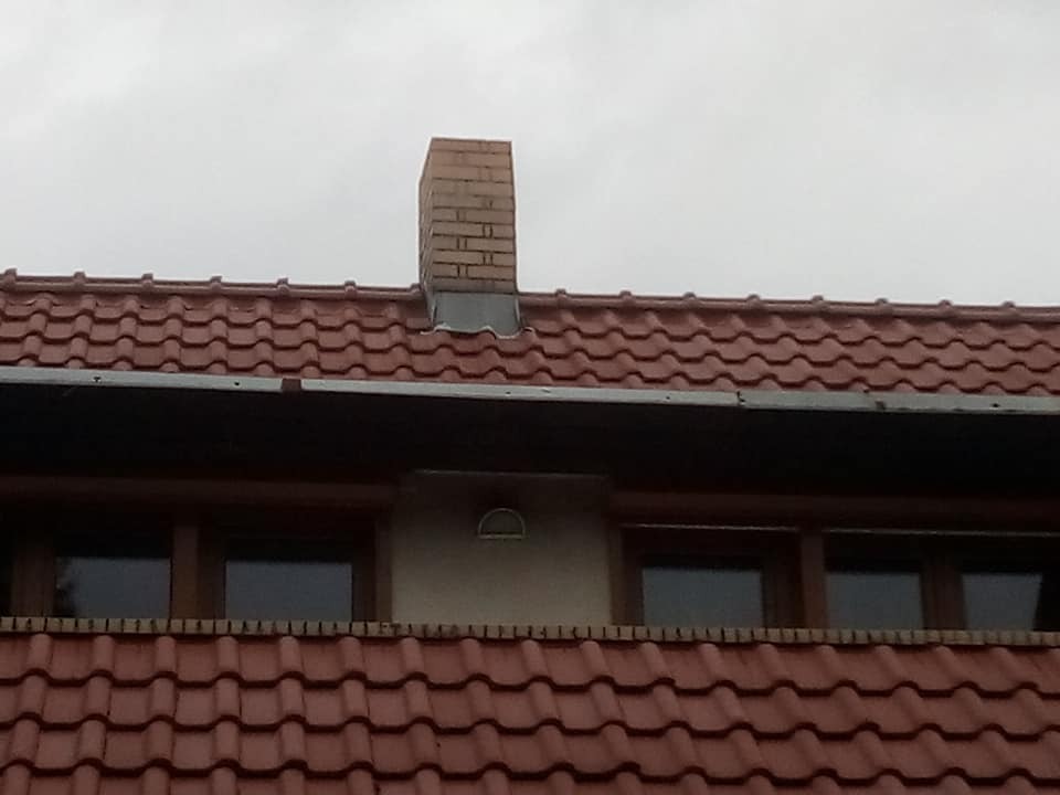 Czerwona dachówka ceramiczna z kominem z cegły, widoczne okno dachowe i rynna, pochmurne niebo w tle.