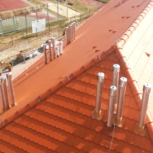 Czerwony dach z dachówki ceramicznej widziany z góry, z licznymi kominami ze stali nierdzewnej, w tle boisko sportowe.