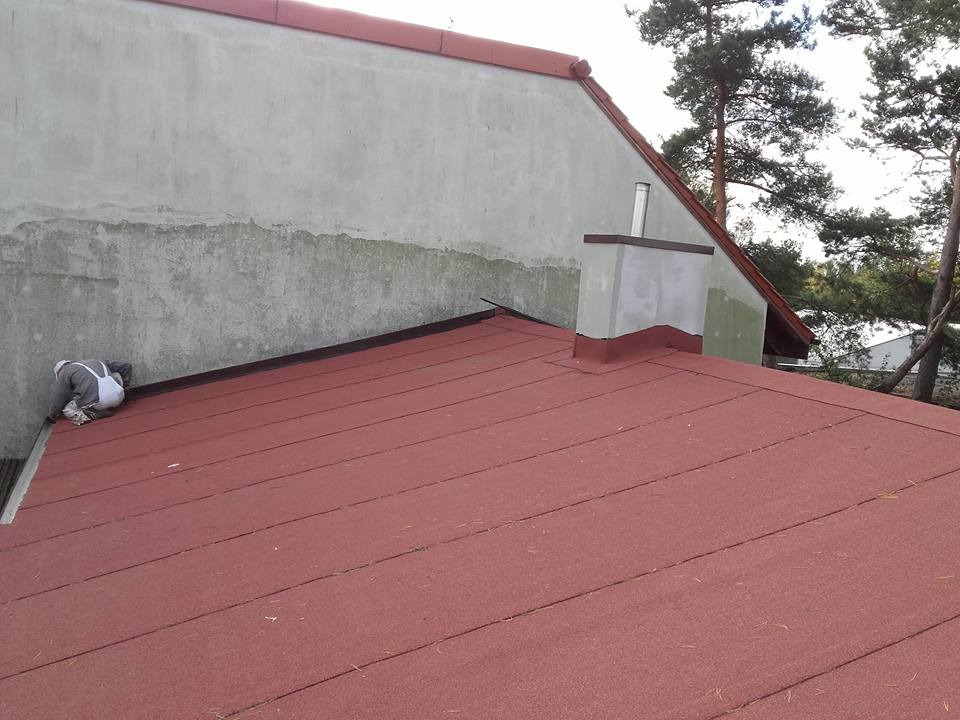 Pracownik w kombinezonie naprawia brzeg dachu pokrytego czerwoną papą bitumiczną, widoczny komin i ściana budynku z wyraźnymi śladami wilgoci.