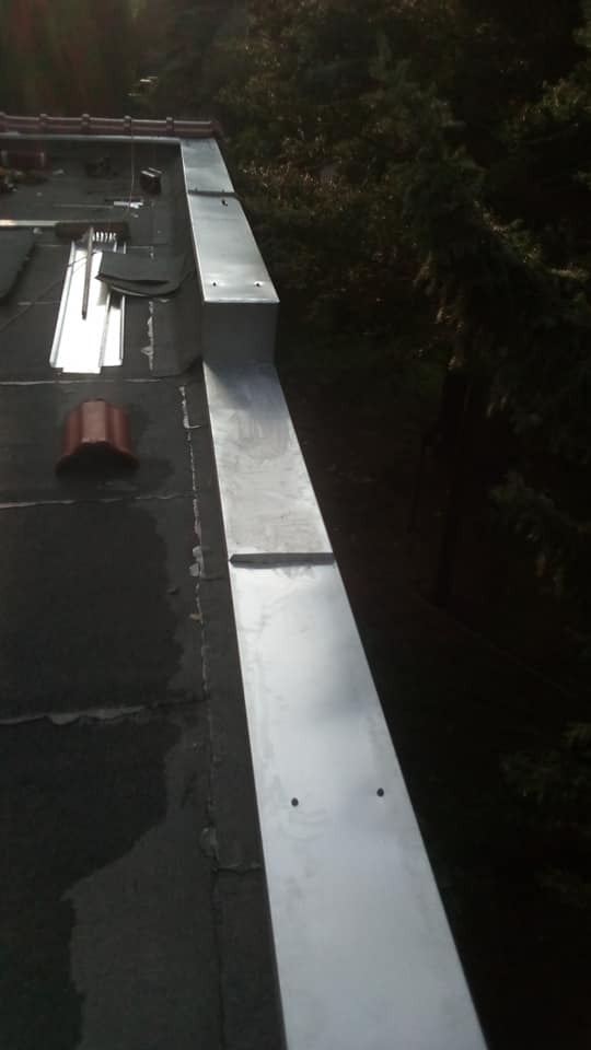 Świeżo zamontowana, długa, metalowa obróbka blacharska na krawędzi płaskiego dachu pokrytego papą; widoczne narzędzia i elementy dekarskie.