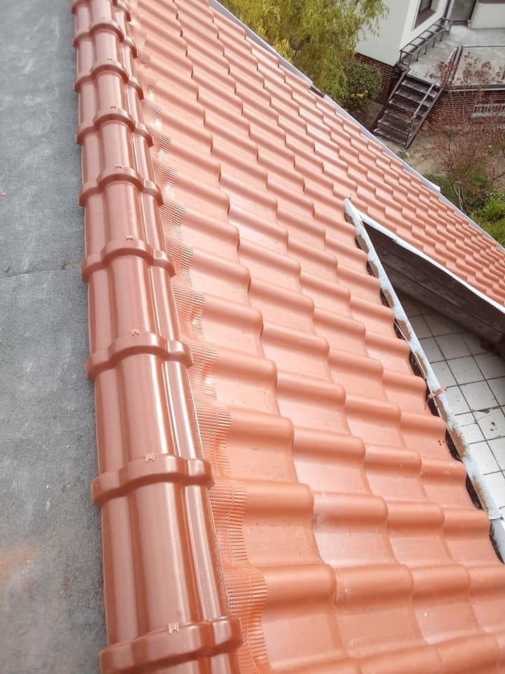 Czerwona dachówka ceramiczna ułożona na dachu domu, widoczna rynna i fragment okna dachowego, tło stanowią drzewa i część budynku.