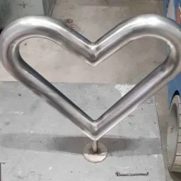 Metalowa poręcz w kształcie serca, wykonana ze stali nierdzewnej, zamocowana na okrągłej podstawie, widoczna na tle podłogi warsztatowej i niebieskiego wózka widłowego.